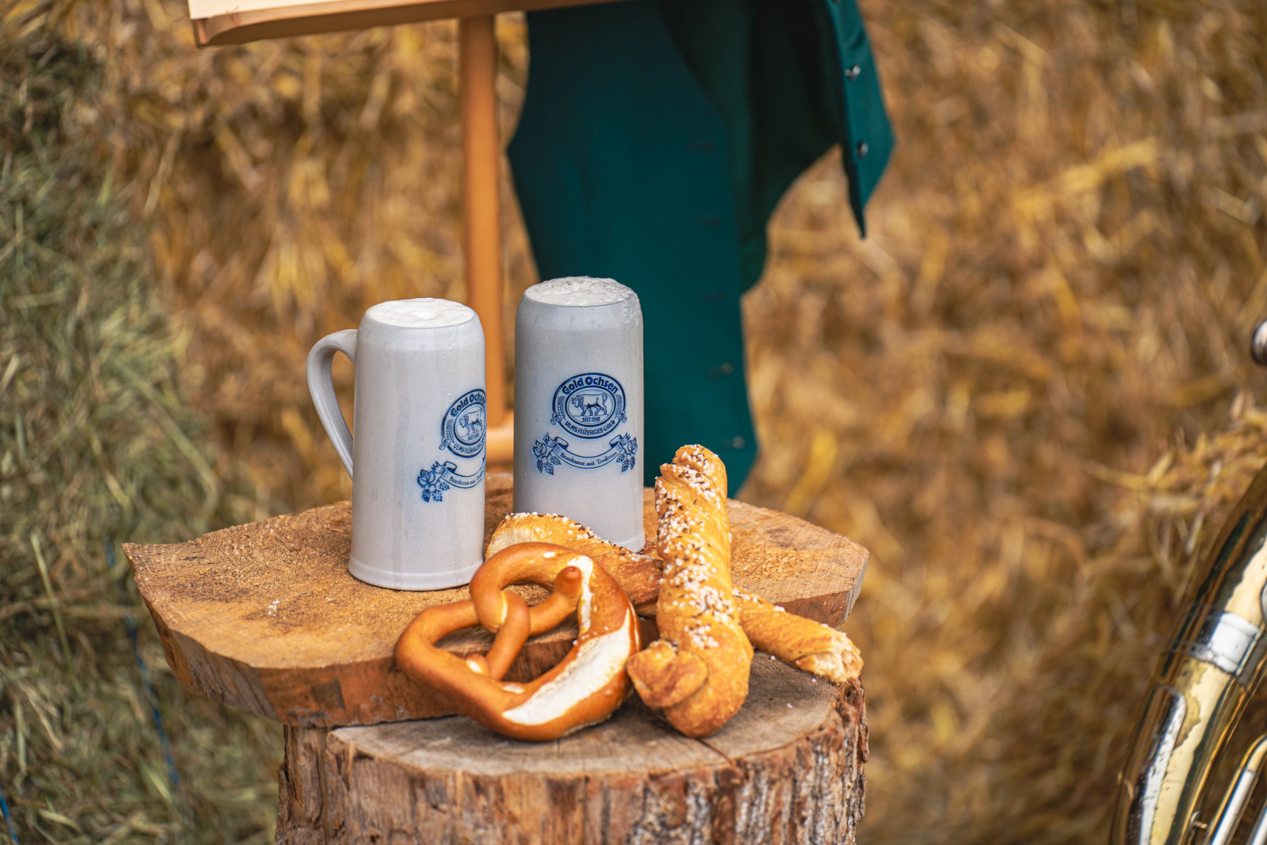 Zwei Steinkrüge mit Bier stehen auf einem Holzstamm. Davor liegen eine Brezel und eine Seele.